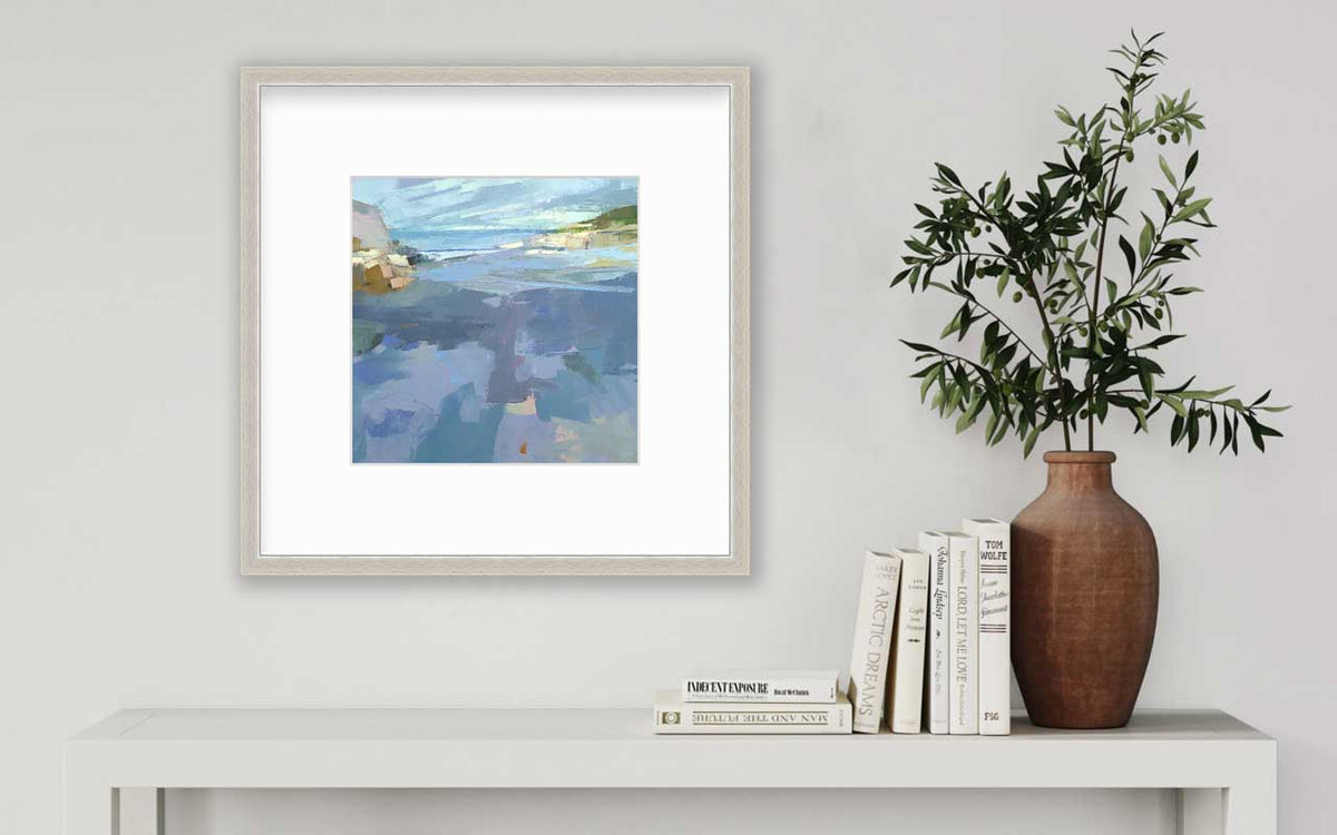 A blue, purple and green abstract landscape print in a silver frame with a mat hangs on a white wall above a white side table with a stack of books and a terracotta vase with greenery stems.
