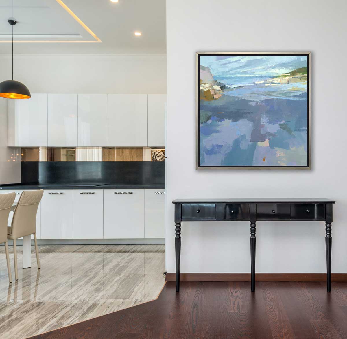 A blue, purple and green abstract landscape print in a silver floater frame hangs on a white wall above a black console table, next to an opening to a kitchen with white cabinets, black countertops and two beige chairs.