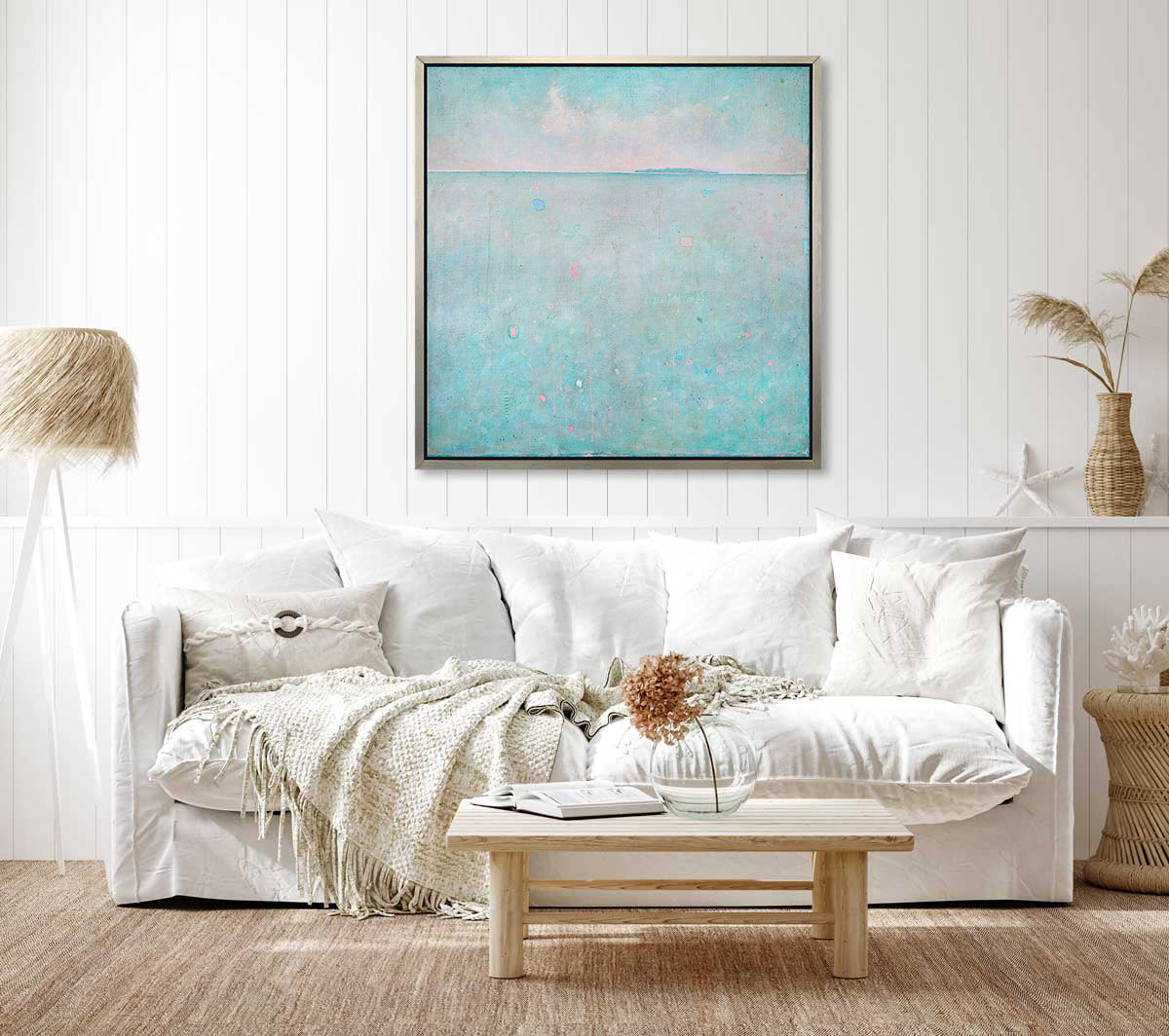 A light blue abstract landscape print in a silver floater frame hangs on a white wall above a white sofa with matching pillows, a low wooden coffee table and a white tripod floor lamp.
