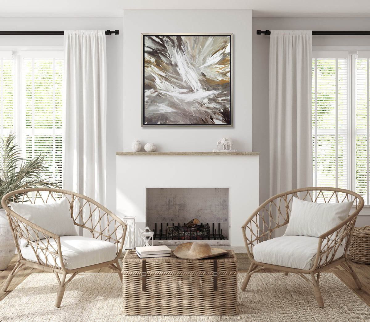 A white, grey, dark grey and gold abstract print in a silver floater frame hangs on a white wall above a white fireplace, two woven chairs with white cushions and a wicker trunk.