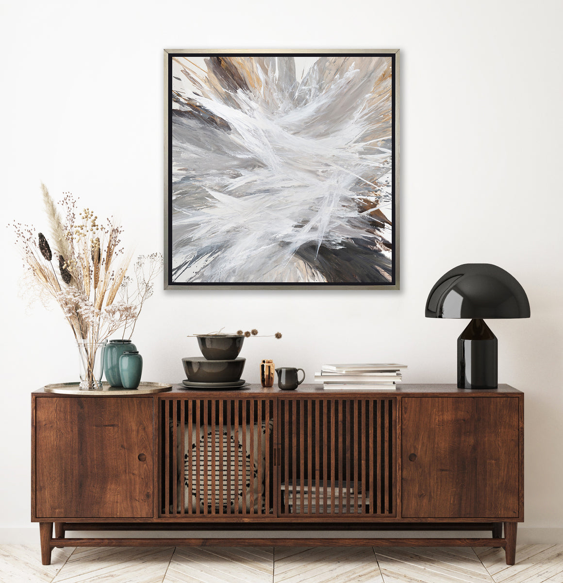 A white, grey, dark grey and gold abstract print in a silver floater frame hangs on a white wall above a dark wood credenza with a black modern table lamp, a stack of books, a stack of bowls and a several vases with wheat stems.