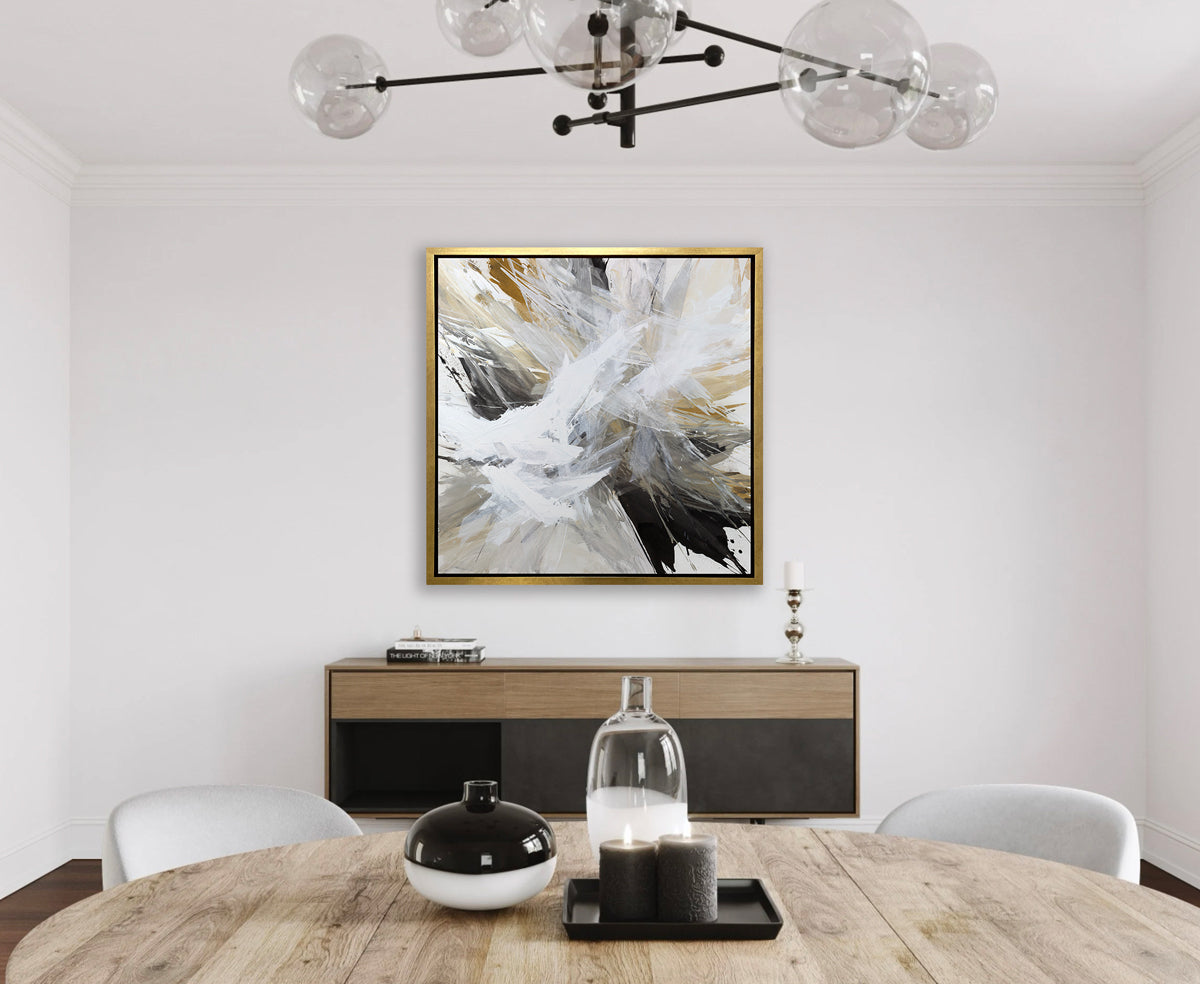 A white, black, grey and gold abstract print in a gold floater frame hangs on a white wall above a black and dark wood sideboard, a round wood table and under a modern black iron light fixture.