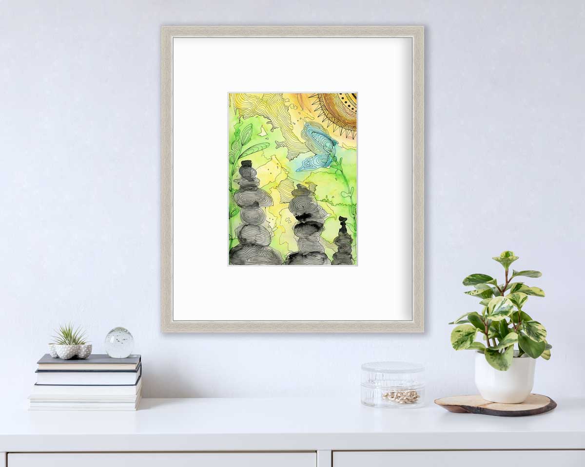 A green, yellow, and black illustration print in a silver frame with a mat hangs on a white wall above a white surface with a stack of books, a clear round container with gold accessories and a potted plant.