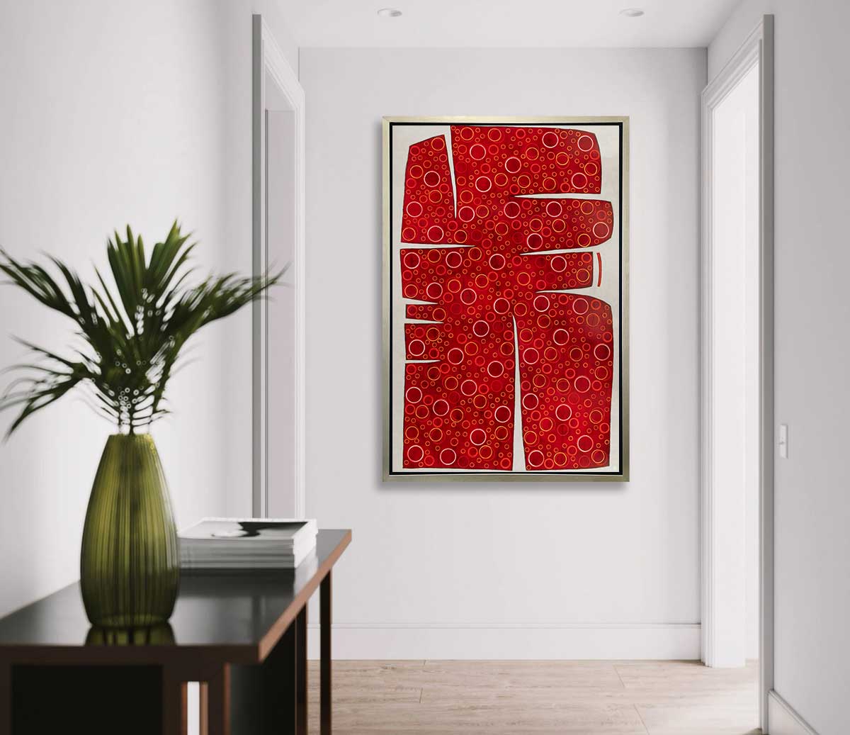A red, yellow and white abstract print in a silver floater frame hangs on a white wall at the end of a hallway across from a black console table with a stack of books and a green vase with greenery stems.