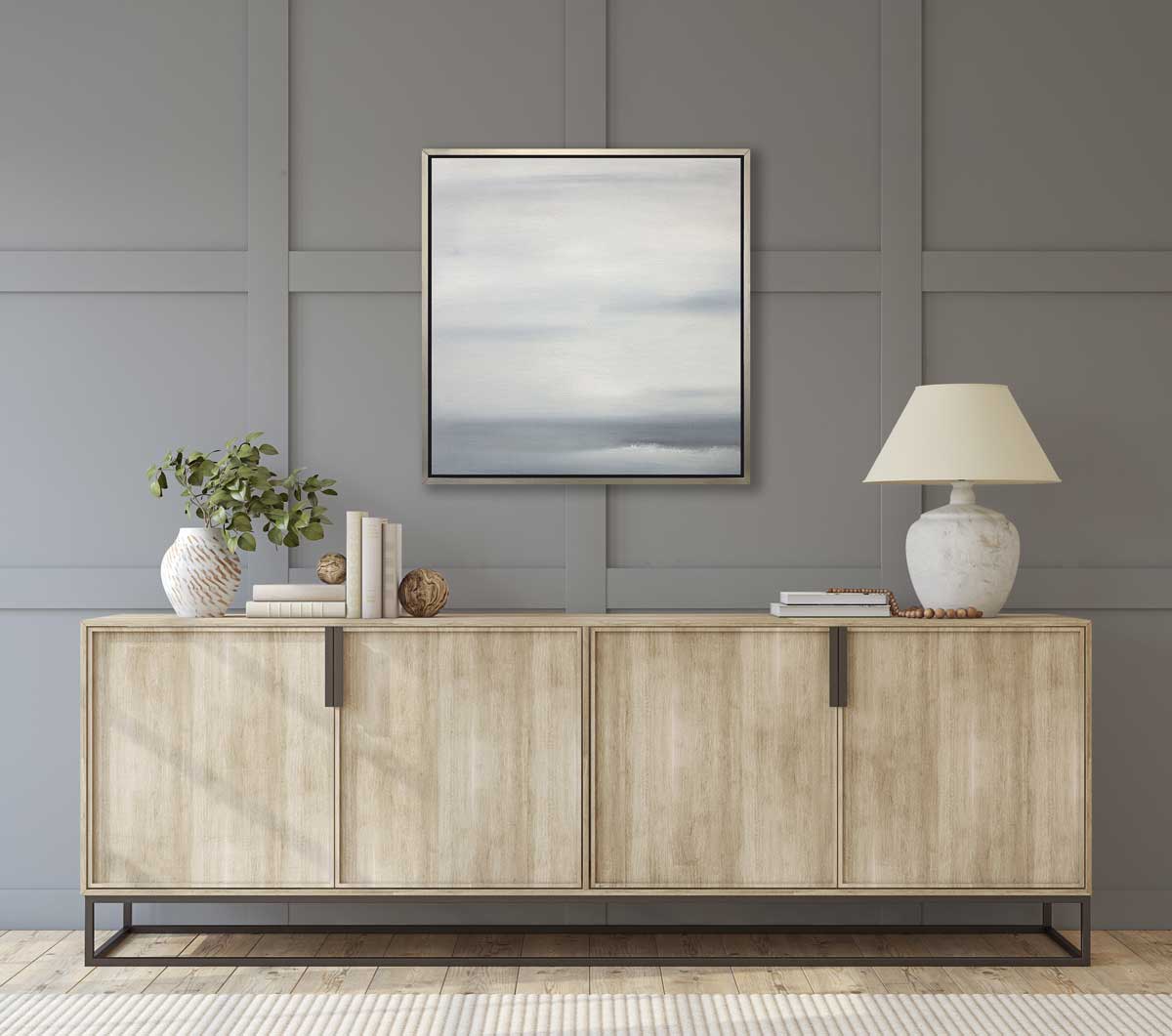 A tonal grey print in a silver floater frame hangs on a grey window-paned wall above a natural wood sideboard with a table lamp, stacks of books and a white vase with eucalyptus stems.
