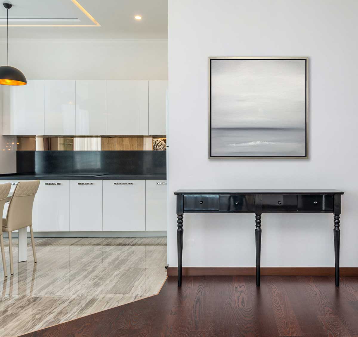 A tonal grey print in a silver floater frame hangs on a white wall above a black console table and an opening to a kitchen with white cabinets, black counters and beige chairs.