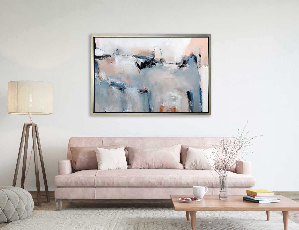 A blue, light pink and white abstract print in a silver floater frame hangs on a white wall above a soft pink sofa with pillows, a rectangular wood coffee table and a tall floor lamp.