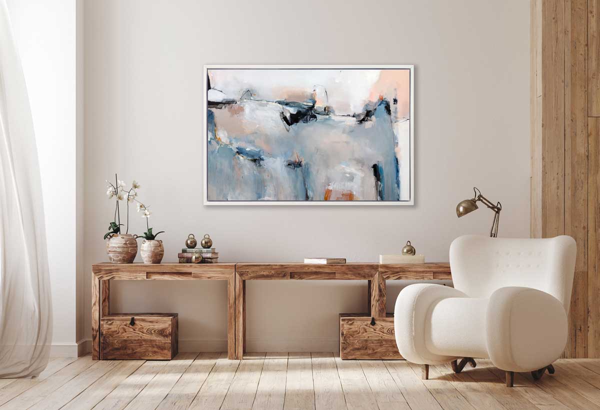 A blue, light pink and white abstract print in a white floater frame hangs on a white wall above a long natural wood desk with accessories, two potted white orchids and a white modern chair.