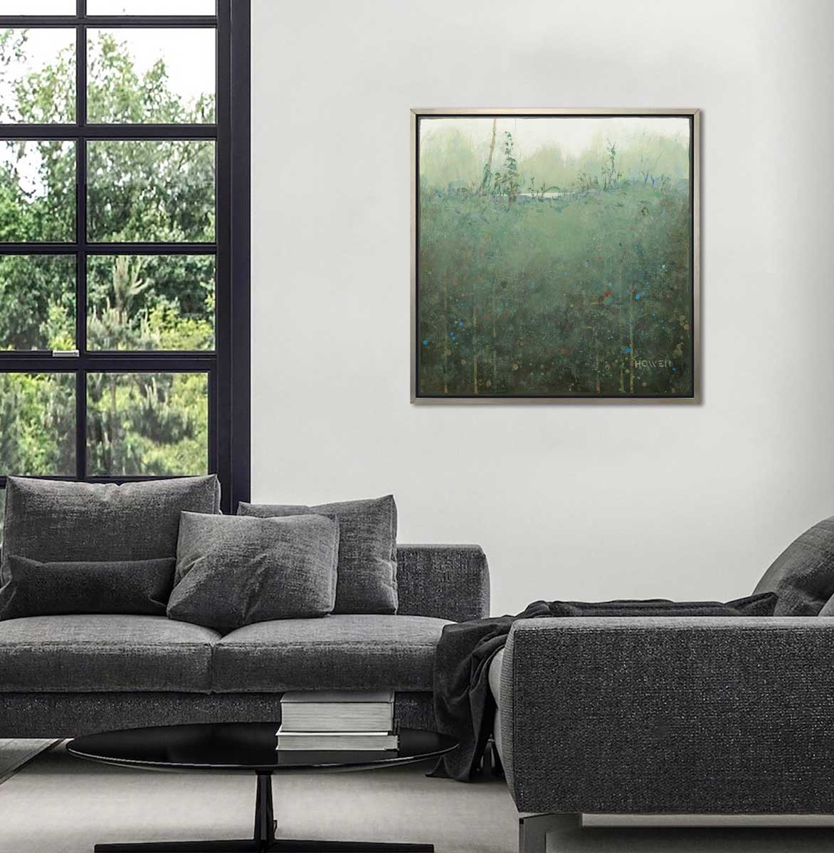A dark muted green abstract landscape print in a silver floater frame hangs on a white wall above a dark grey sofa and chair set and a round black coffee table.