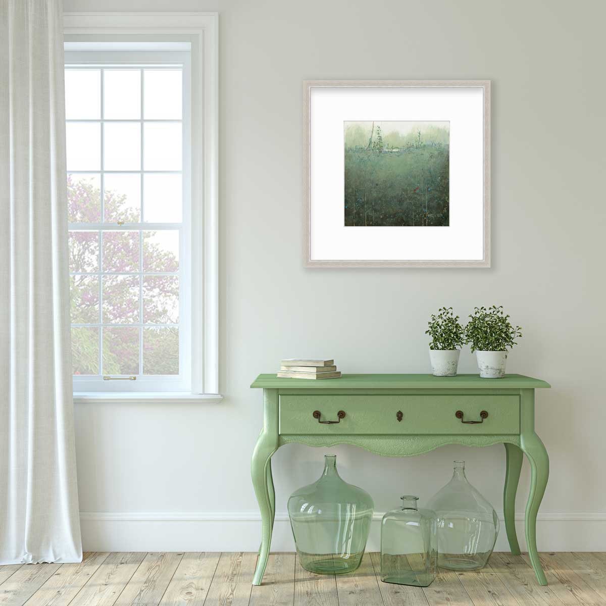 A dark muted green abstract landscape print in a silver frame with a mat hangs on a white wall above a green console table with a stack of books, two potted plants and three green glass bottles.