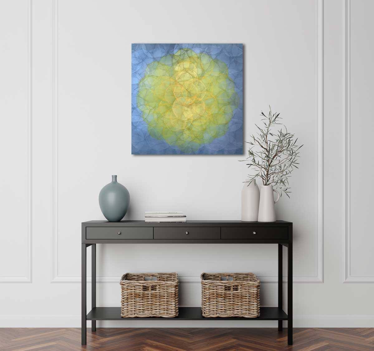 A blue and yellow abstract geometric painting hung on a white wall above a black console table. Atop of the table sits a blue vase, a stack of books, and two white ceramic vases with flowers.