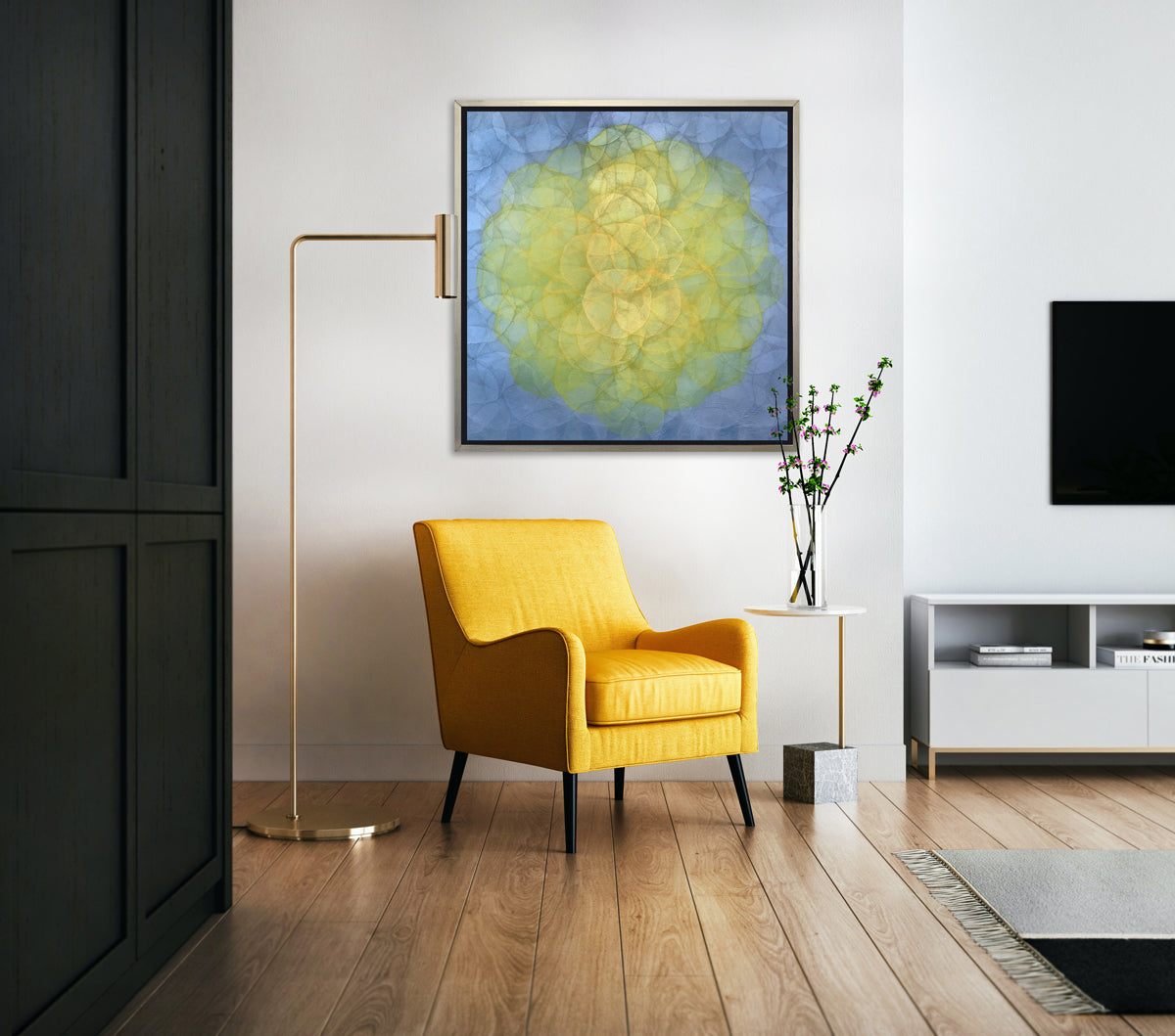 A blue and yellow abstract geometric print in a silver floater frame hangs on a white wall above a deep yellow chair, a tall gold floor lamp and a small side table with a glass vase with purple and green stems.