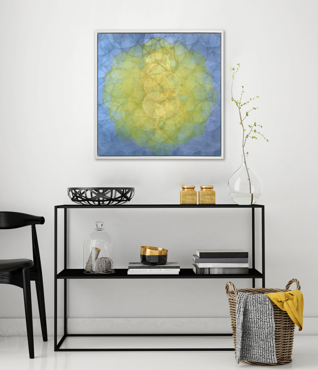 A blue and yellow abstract geometric print in a white floater frame hangs on a white wall above a black two tier shelf with stacks of books, a black decorative bowl, two gold containers and a glass vase with a greenery stem.