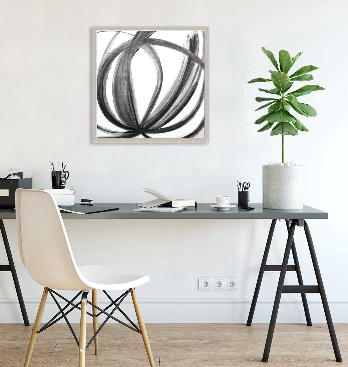 A black, grey and white abstract print in an unmatted silver frame hangs on a white wall above a black desk with open notebooks, a black mug with pencils and a tall potted plant.