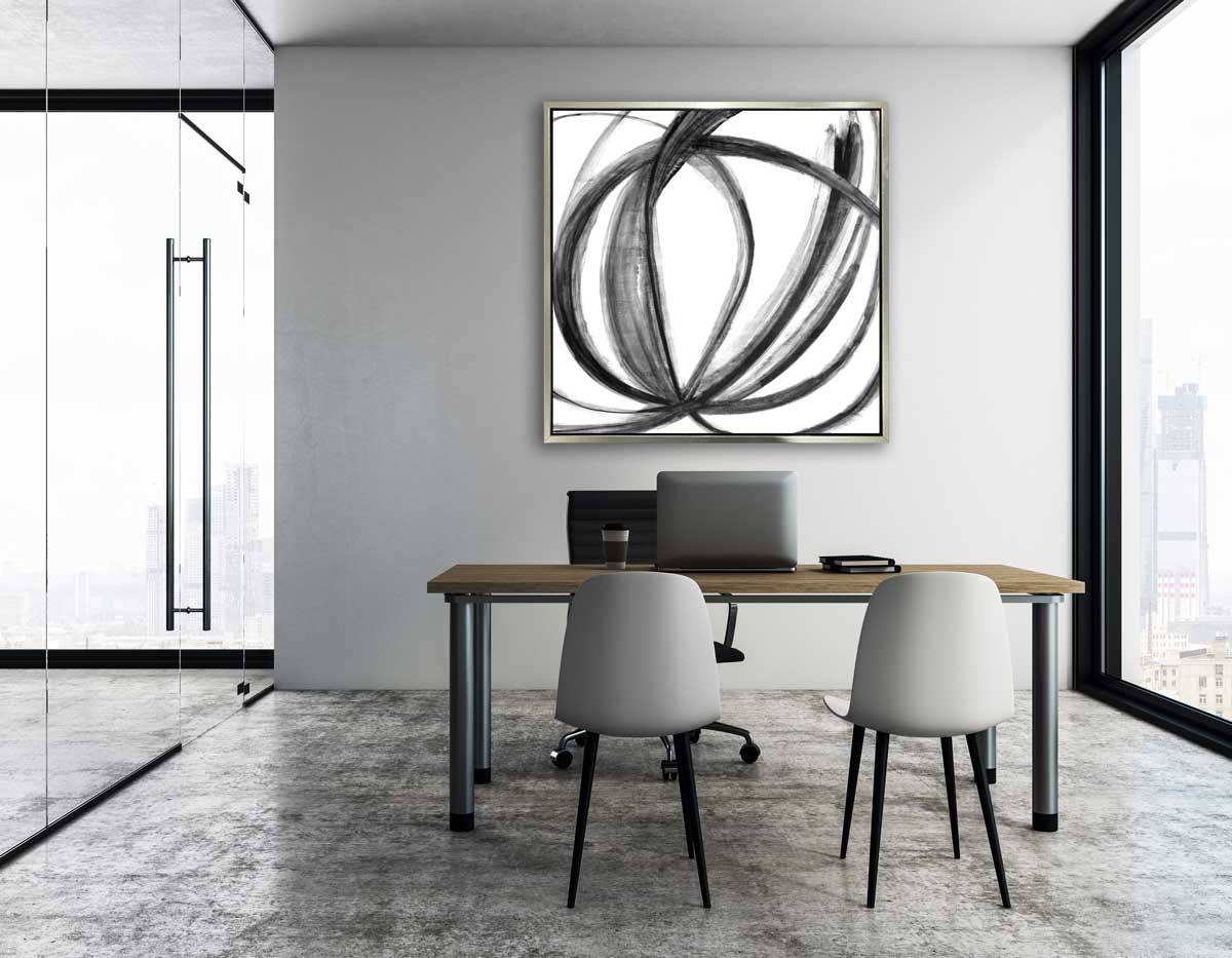 A black and white abstract print in a silver floater frame hangs on a grey wall above a desk with a laptop computer, one black chair and two white chairs.