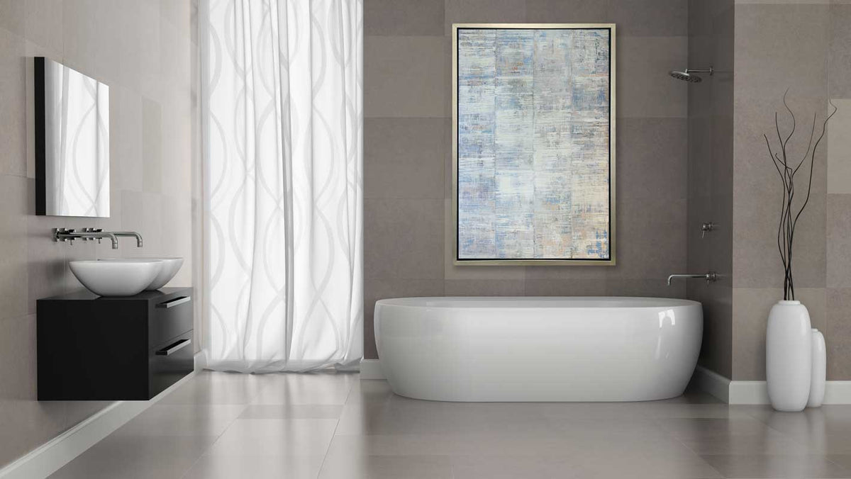 A white and blue abstract print in a silver floater frame hangs on a grey wall above a white bathtub.