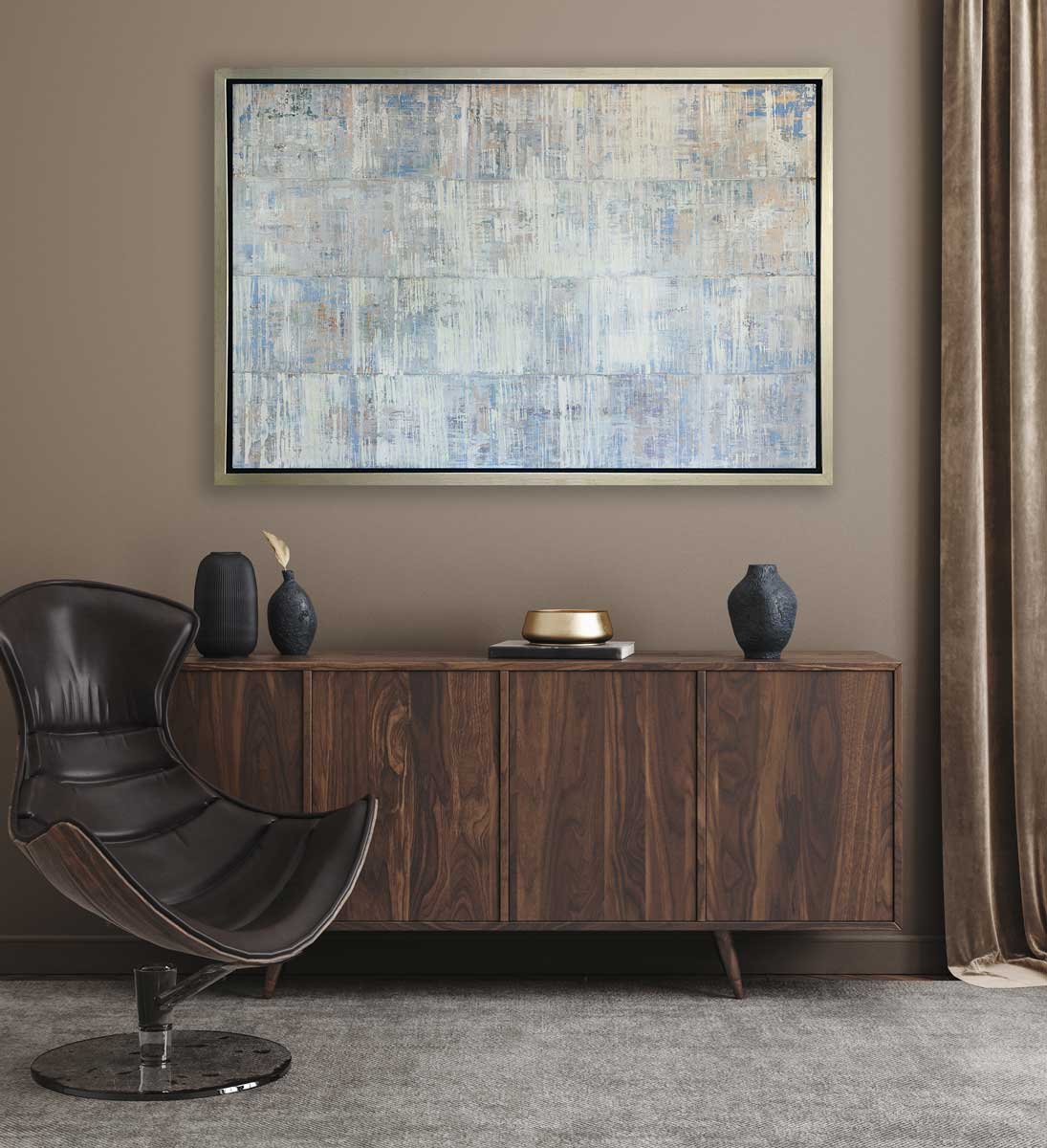 A white and blue abstract print in a silver floater frame hangs horizontally on a beige wall above a wooden sideboard with black vases and a brown modern chair.