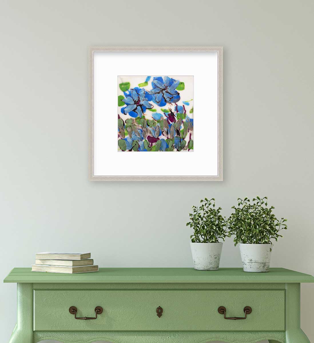 A blue, green and white abstract floral print in a silver frame with a mat hangs on a white wall above a green console table with a stack of books and two potted plants.