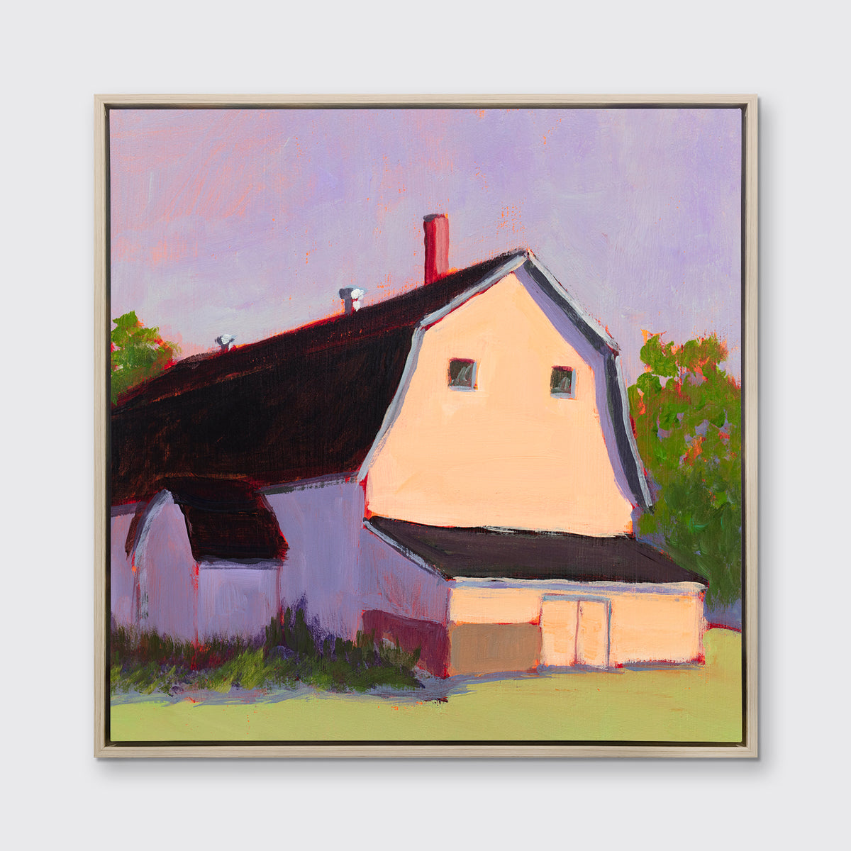 A print of a beige barn, a lavender sky, green bushes and trees and a light green foreground in a white oak wood floater frame hangs on a white wall.