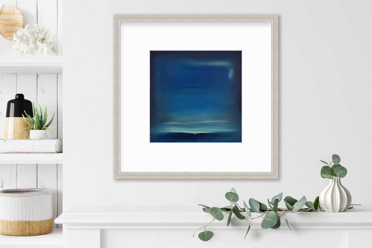 A dark blue and teal with accents of light yellow abstract print in a silver frame with a mat hangs on a white wall above a white surface with a eucalyptus filled white vase and next to a built in bookshelf with decorative accessories.