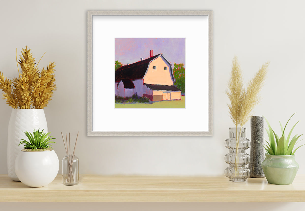A print of a beige barn, a lavender sky, green bushes and trees and a light green foreground in a silver frame with a mat hangs on a white wall above a light wood surface with several vases with wheat stems and greenery.