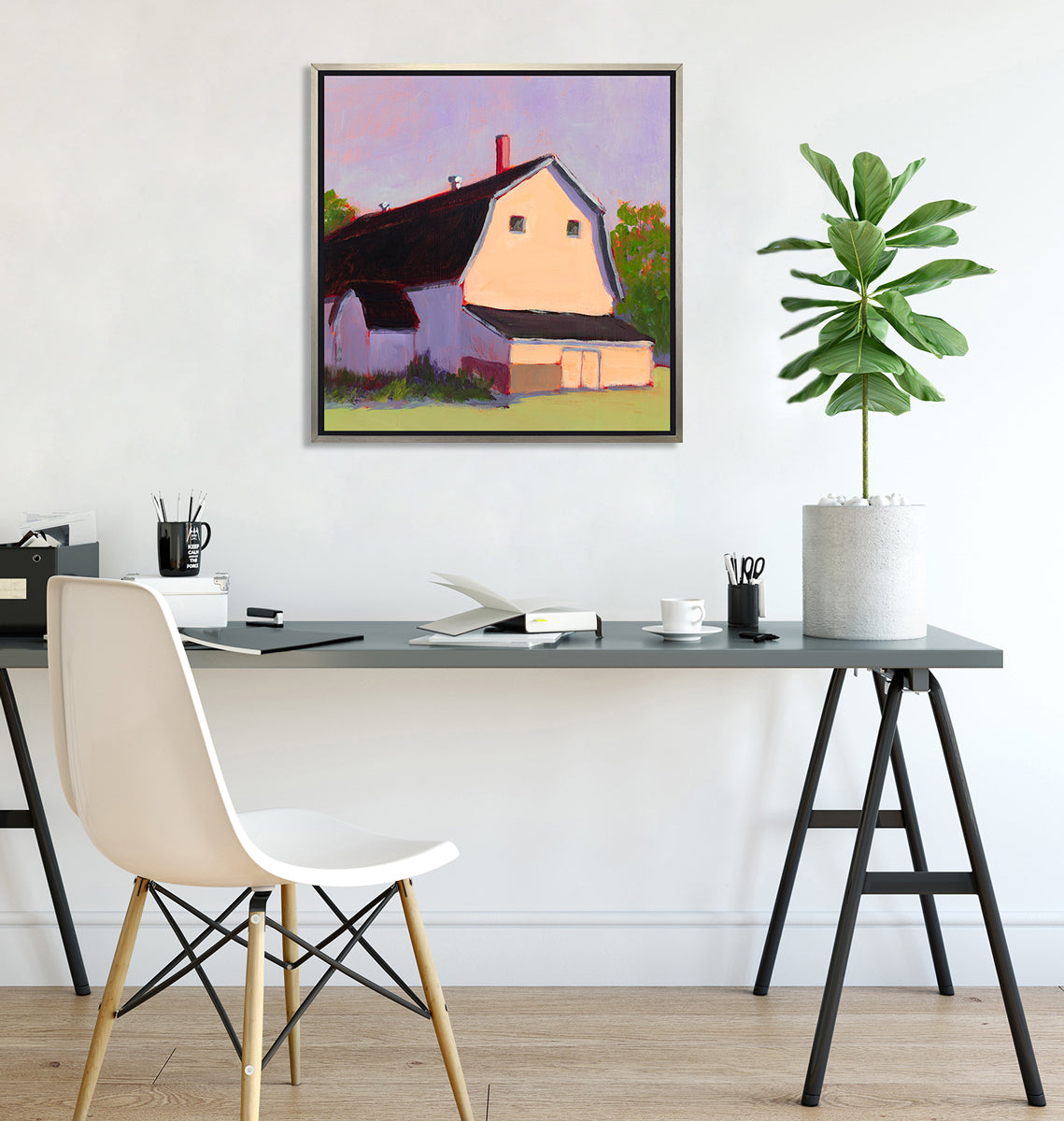 A print of a beige barn, a lavender sky, green bushes and trees and a light green foreground in a silver floater frame hangs on a white wall above a grey desk with open notebooks, black mugs with pencils and a potted plant.