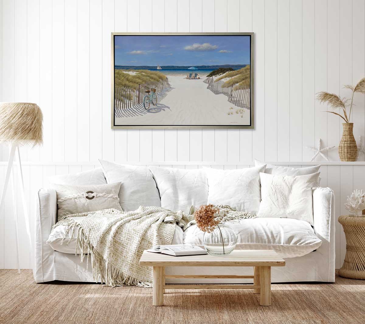 A blue, beige and light green contemporary seascape print in a silver floater frame hangs on a white paneled wall above a white sofa with pillows, a wood coffee table and a tripod floor lamp.