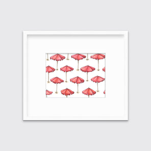 A print of red beach umbrellas with brown poles in a white frame with a mat hangs on a white wall. 