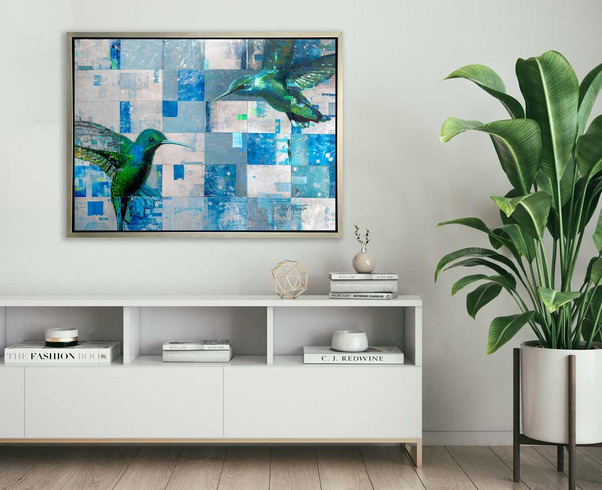 A blue and green abstract hummingbird print in a silver floater frame hangs on a white wall above a white credenza with books, a gold wire sphere, a small vase with two stems and a potted floor plant.