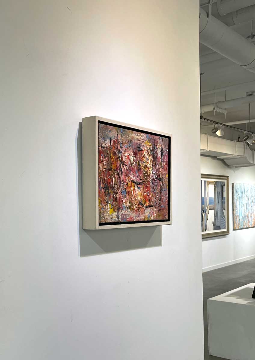 A left side-angle view of a multicolored abstract painting in a white floater frame hangs on a white wall.