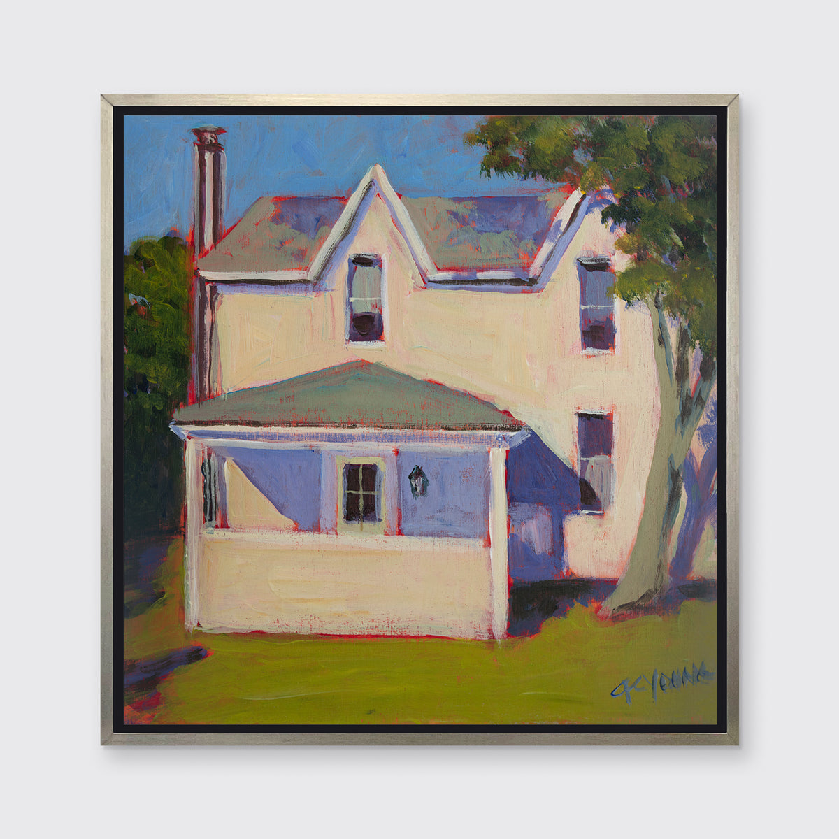 A print of a white house that sits next to a giant tree, green grass in front in a silver floater frame hangs on a white wall.