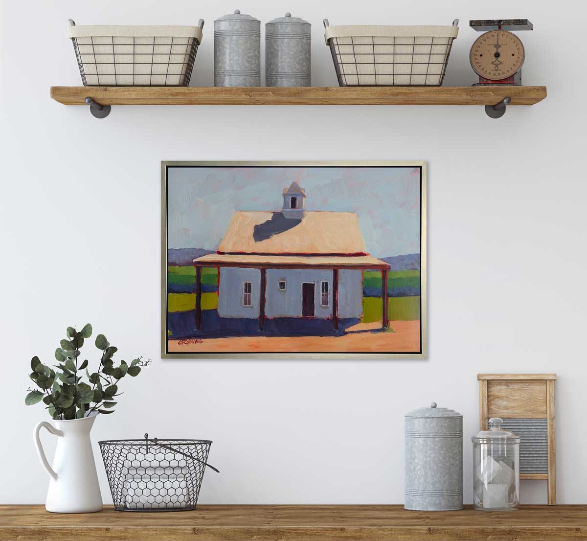 A blue, beige, lavender and green contemporary barn print in a silver floater frame hangs on a white wall underneath a floating shelf with wire baskets and containers and above a shelf with a white pitcher, a wire basket and more containers.