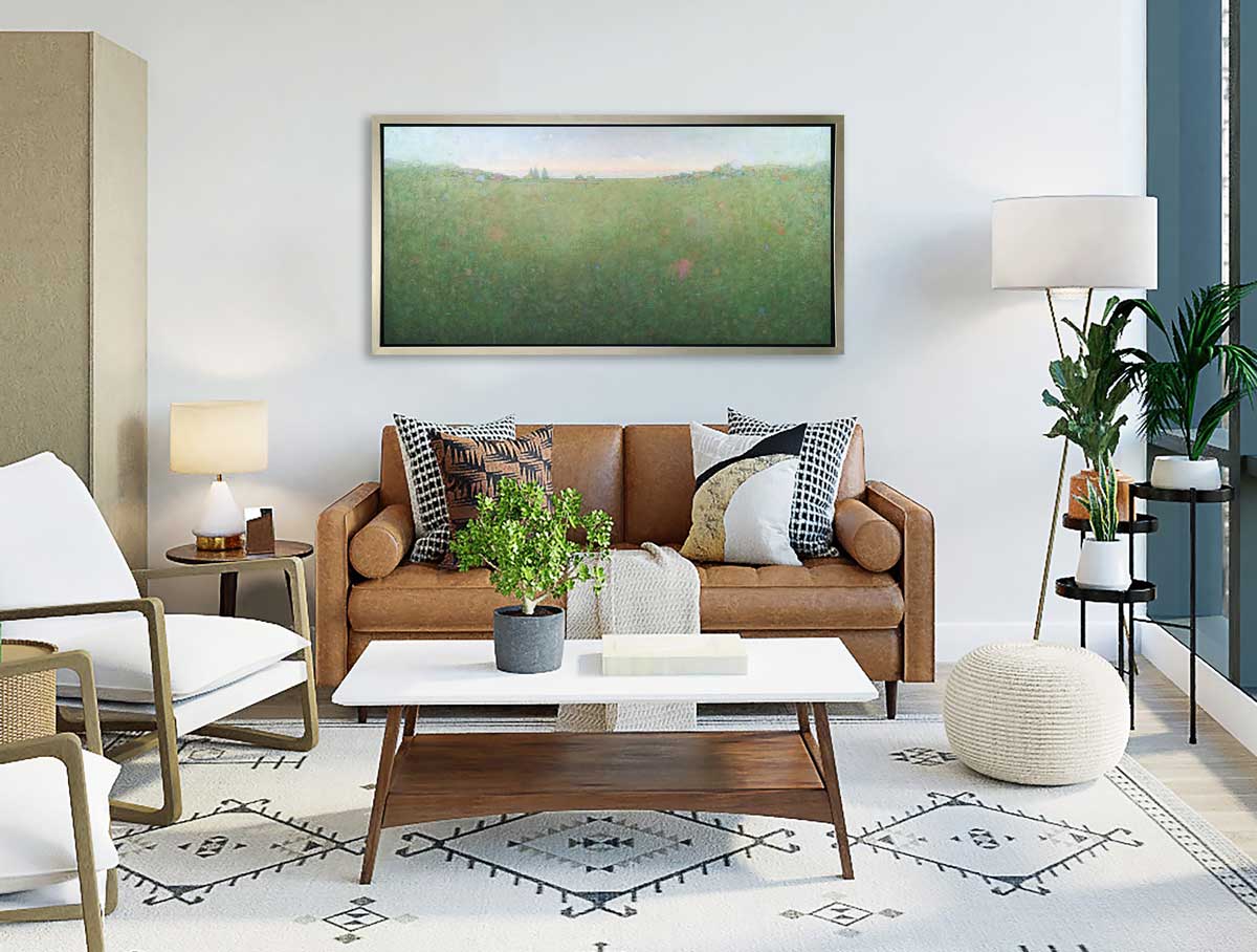 A green abstract landscape print in a silver floater frame hangs on a white wall above a brown leather sofa with pillows, a white coffee table with a small potted shrub and two white chairs.