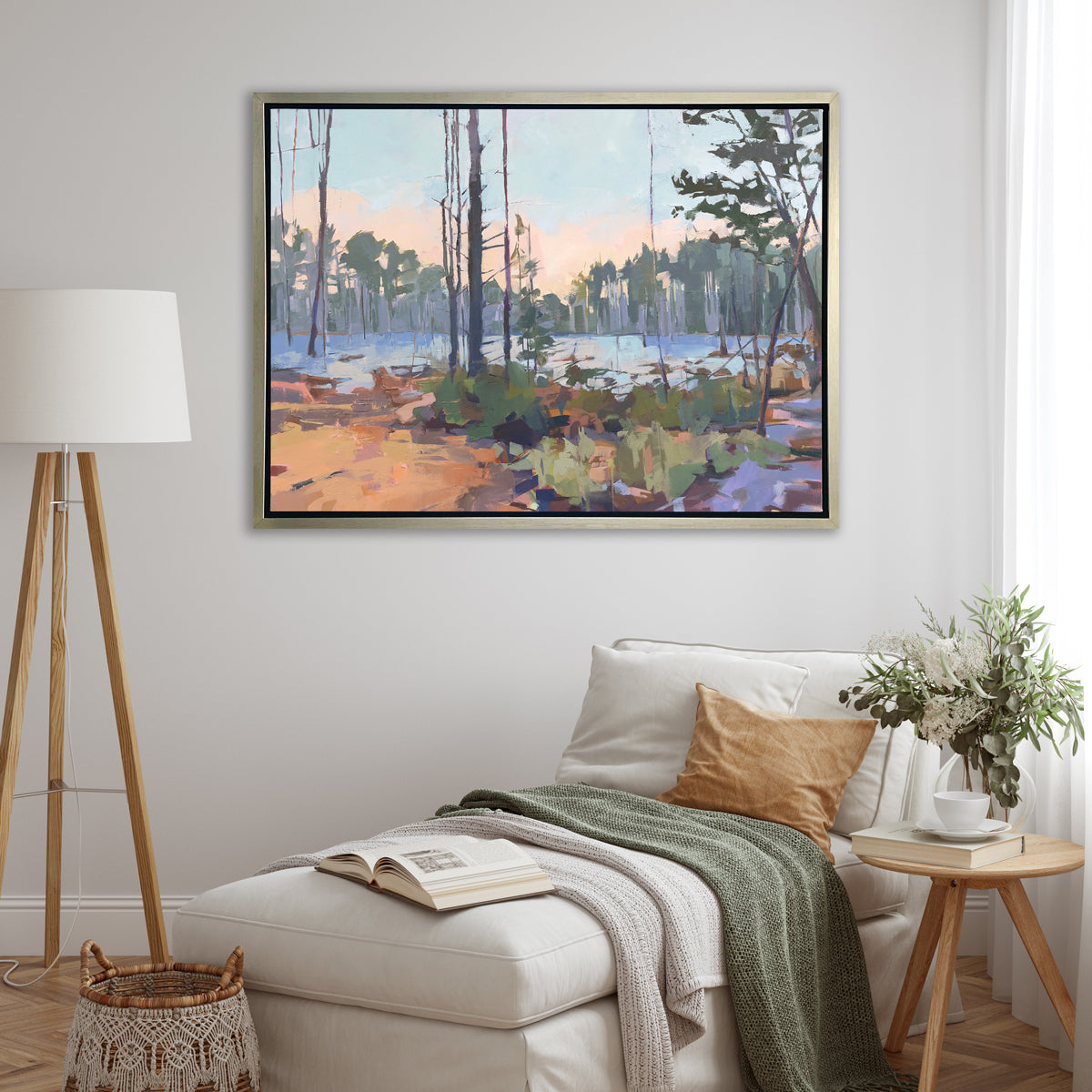 A light blue, green, brown and purple abstract landscape print in a silver floater frame hangs on a white wall above a white lounger chair with blankets and pillows, a wood side table with a glass vase with a floral bouquet and a tall tripod floor lamp.