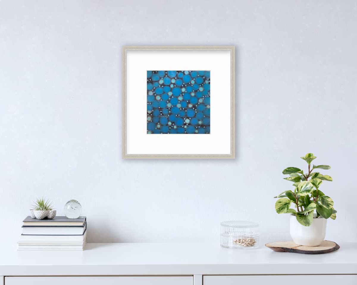 A blue, teal and black abstract circle print in a silver frame with a mat hangs on a white wall above a white surface with a stack of books, a round clear container with gold accessories and a potted plant.