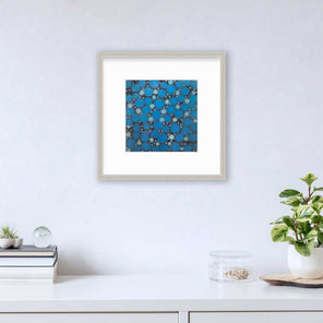 A blue, teal and black abstract circle print in a silver frame with a mat hangs on a white wall above a white surface with a stack of books, a round clear container with gold accessories and a potted plant.