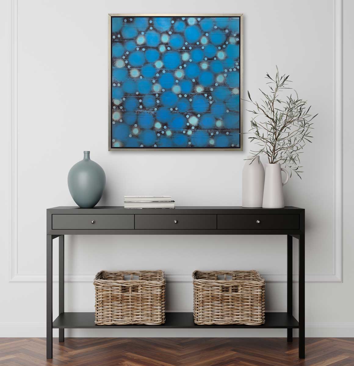 A blue, teal and black abstract circle print in a silver floater frame hangs on a white wall above a black console table with a muted teal vase, two white vases with greenery and two wicker baskets.