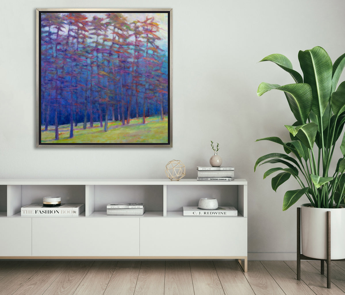 A blue, green, red and pink abstract landscape print in a silver floater frame hangs on a white wall above a white shelf credenza with stacks of books, a gold wire sphere decoration and a large potted floor plant.