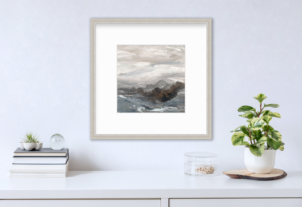 A beige, white, brown and grey abstract print in a silver frame with a mat hangs on a white wall above a white surface with a stack of books, a round clear container with gold accessories and a potted plant.