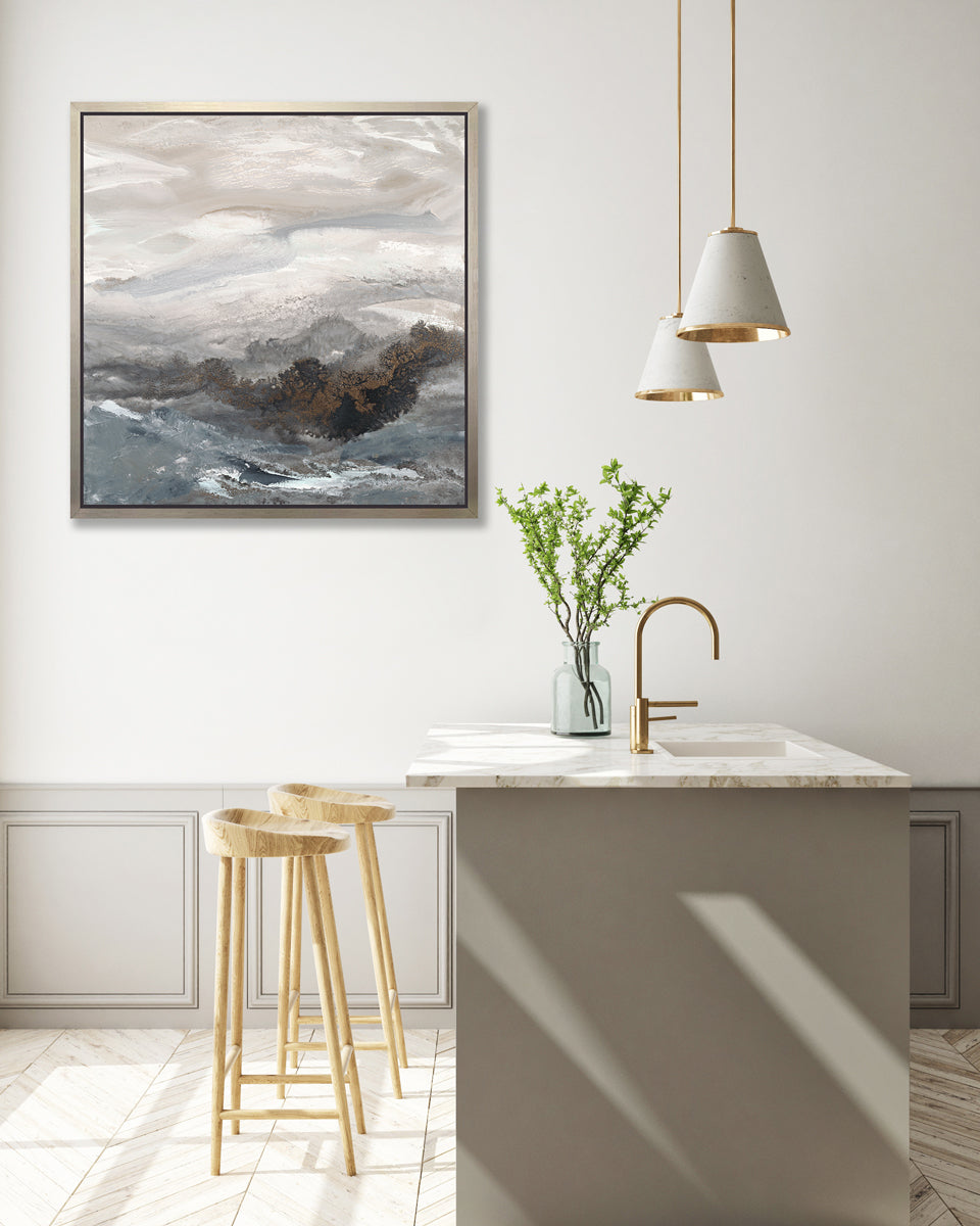A beige, white, brown and grey abstract print in a silver floater frame hangs on a white wall above a white marble kitchen island with a sink and a glass bottle with greenery stems and two wood counter stools.