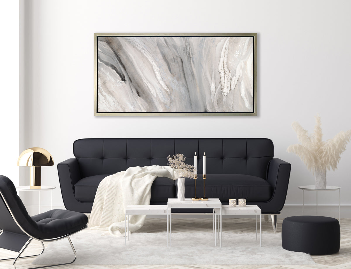 A beige, white and gold abstract print in a silver floater frame hangs on a white wall above a black tufted sofa with a white throw blanket, a black chair, a black ottoman and a white two-tiered coffee table.