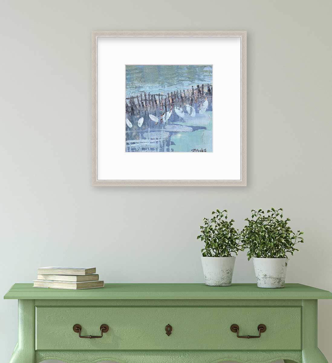 A blue, lavender and olive green abstract print in a silver frame with a mat hangs on a white wall above a green console table with a stack of books and two potted plants.