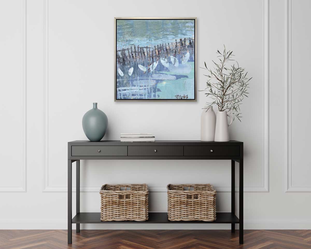 A blue, lavender and olive green abstract print in a silver floater frame above a black console table with a muted teal vase, a stack of books, two white vases with greenery stems and two wicker baskets.