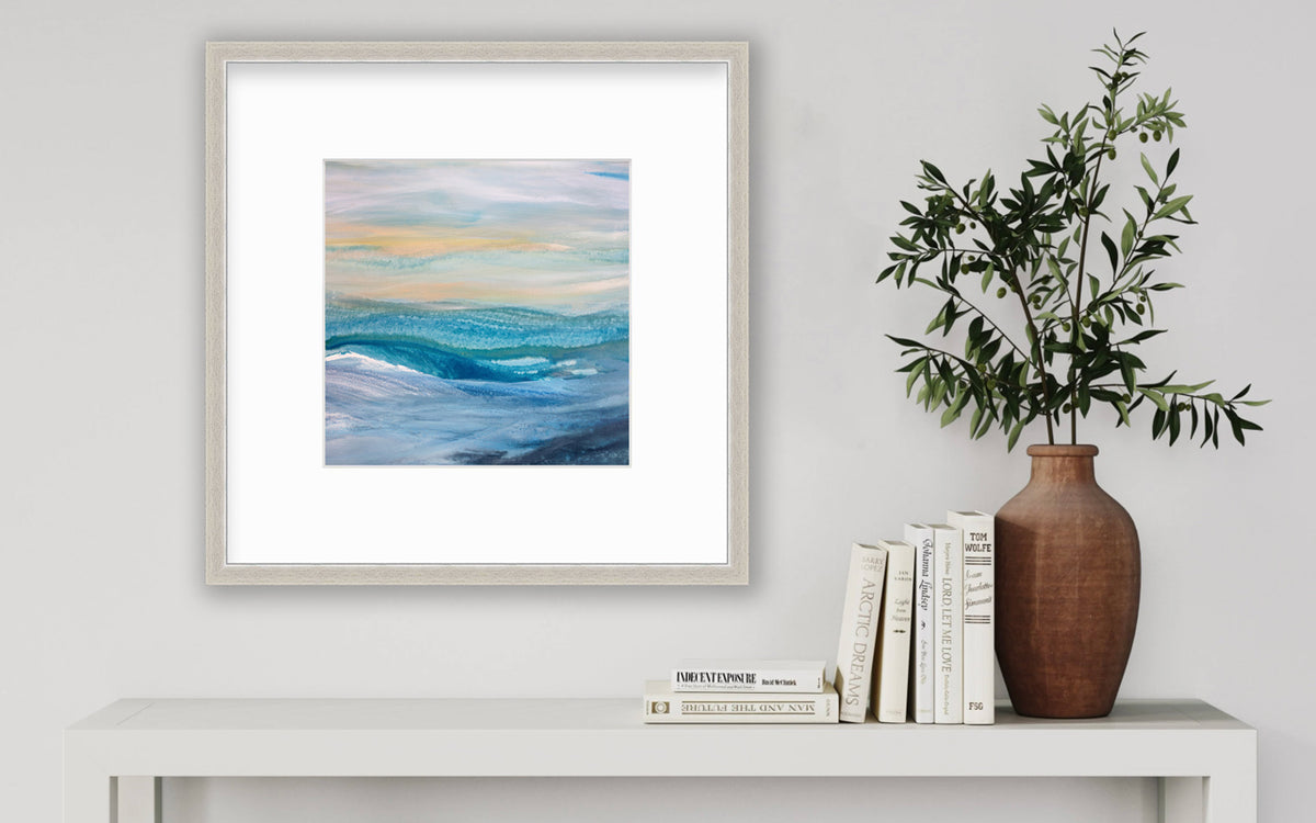 A blue, teal, yellow and white abstract print in a silver frame with a mat hangs on a white wall above a white side table with a stack of books and a terracotta vase with greenery stems.