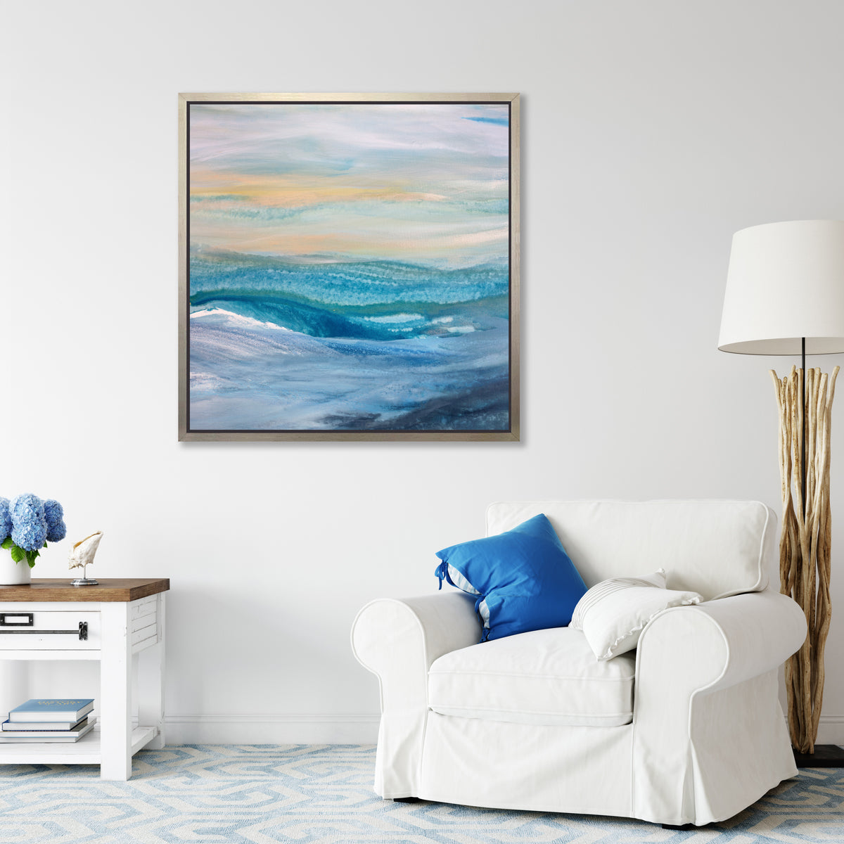 A blue, teal, yellow and white abstract print in a silver floater frame hangs on a white wall above a white oversized chair with pillows, a driftwood floor lamp and a white side table with a stack of books, a blue hydrangea arrangement and a seashell decoration.