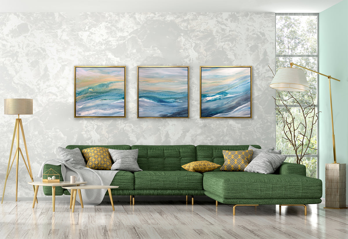 Three blue, teal, yellow and white abstract prints in gold floater frames hang side by side on a white marble wall above a green sectional tufted sofa with yellow and grey pillows, two floor lamps and a two tiered white coffee table.