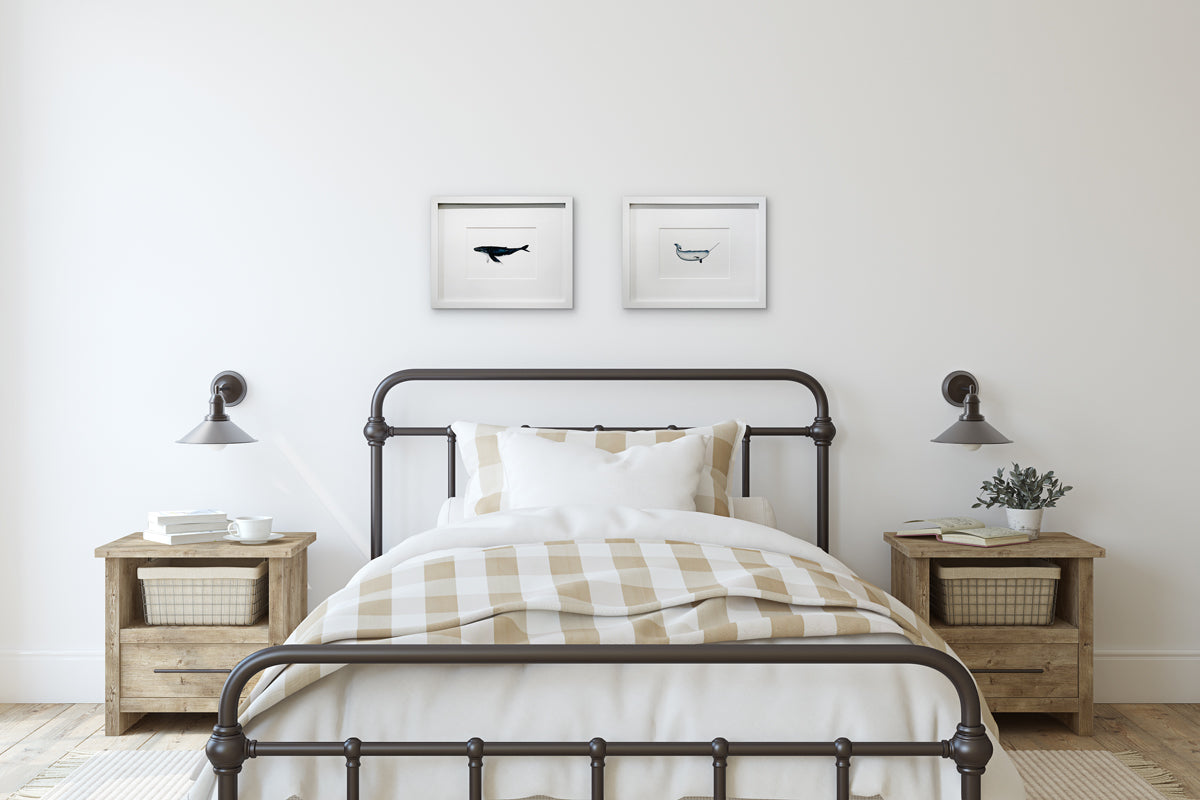 Two illustration drawings of sea creatures in white frames with mats hang on a white wall above a wrought-iron bed with neutral bedding, a set of wood end tables with accessories and two small wall attached pendant lamps.