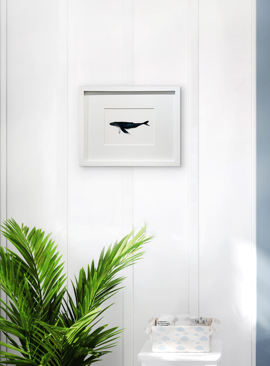 A blue, white and black illustration drawing of a whale in a white frame with a mat hangs on a white wall above a potted floor plant and a white stool with a basket.