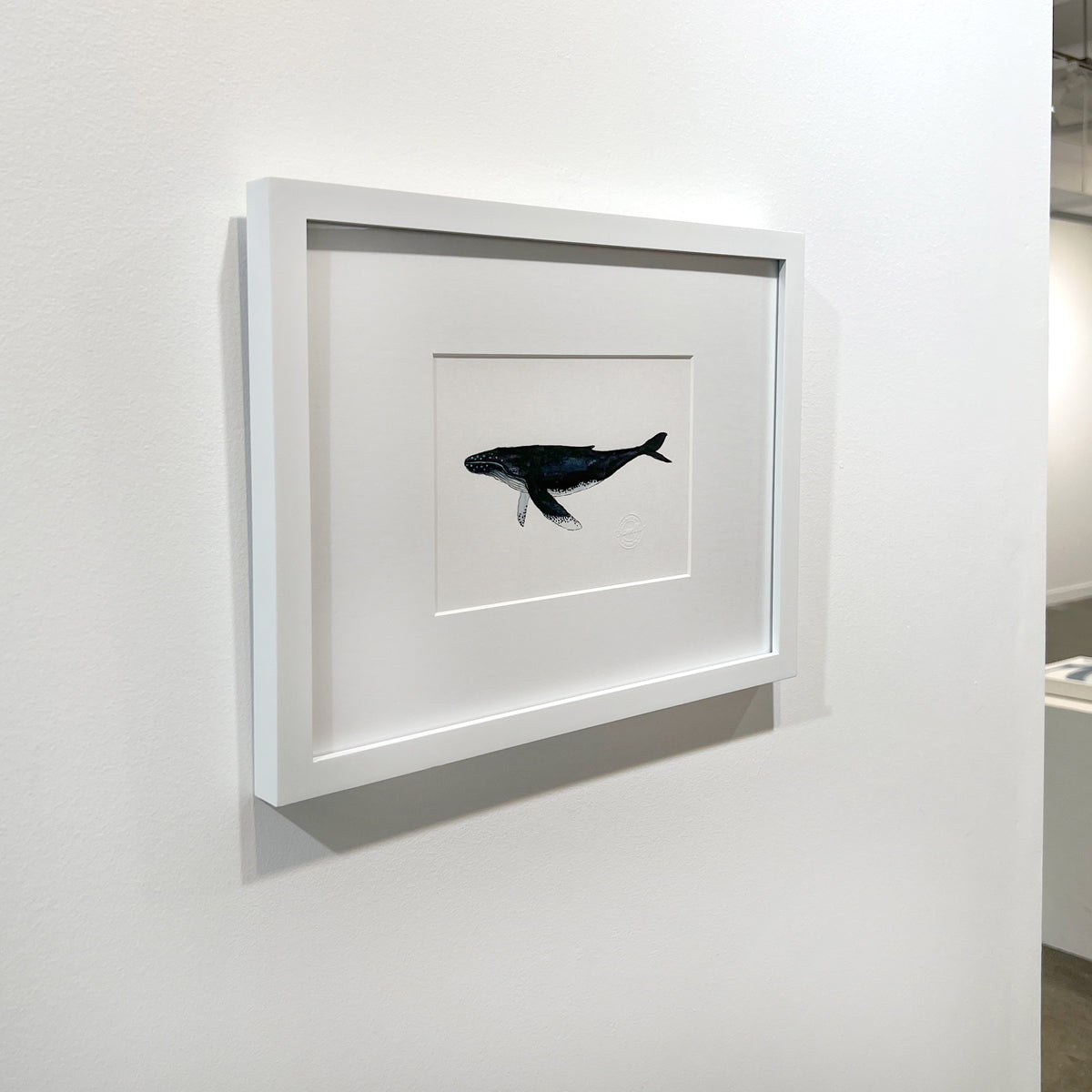 A left side-angle view of a blue, white and black illustration drawing of a whale in a white frame with a mat hangs on a white wall.