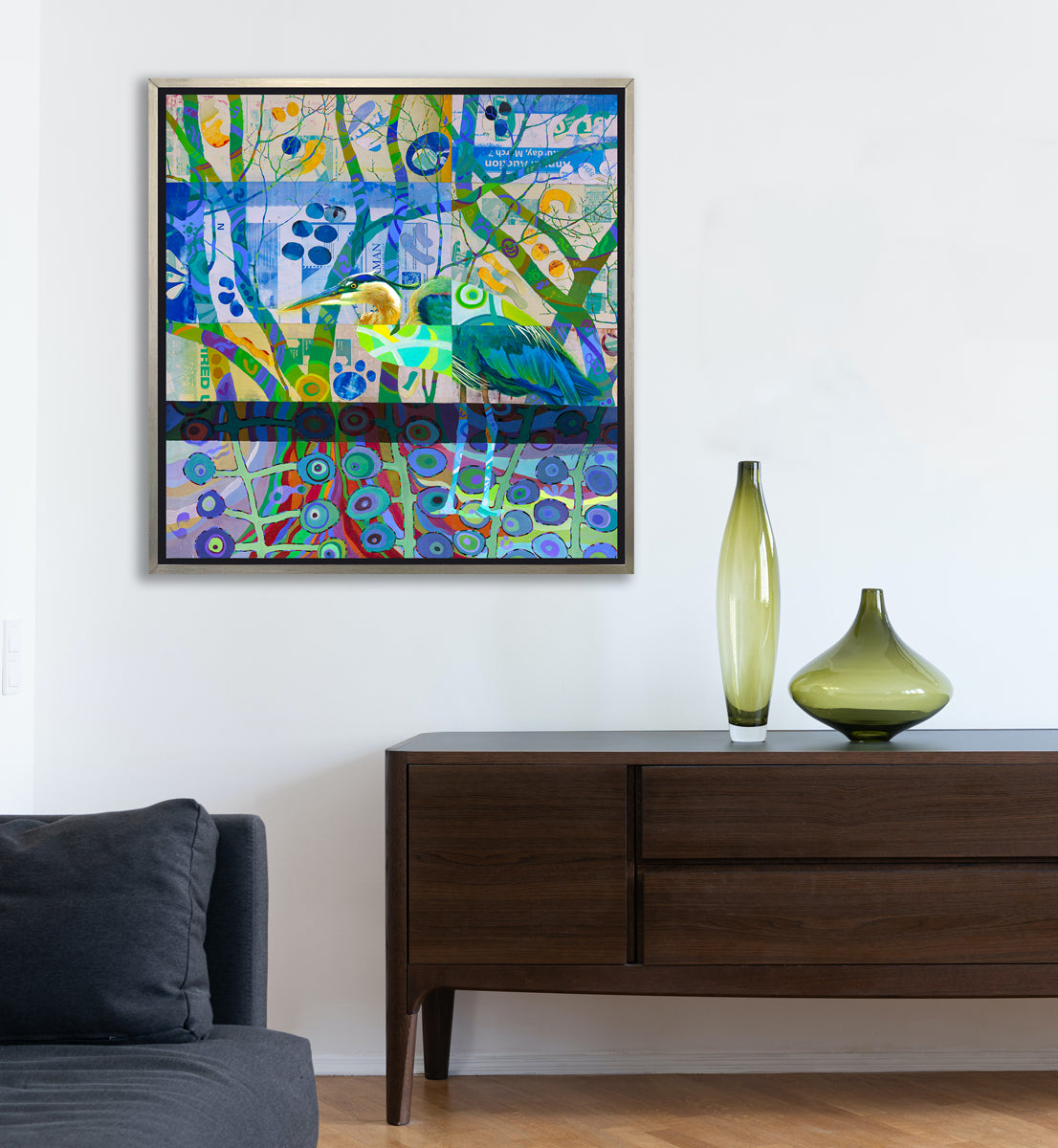 A multicolored abstract print of a bird in a silver floater frame hangs on a white wall above a dark wood console table with two green vases and the edge of a denim blue sofa.